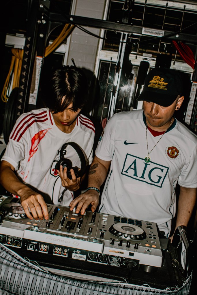 Two DJs mixing music on a digital turntable in a dimly lit environment. One DJ is wearing a white shirt with red accents, while the other wears a white shirt with a logo and a black cap.