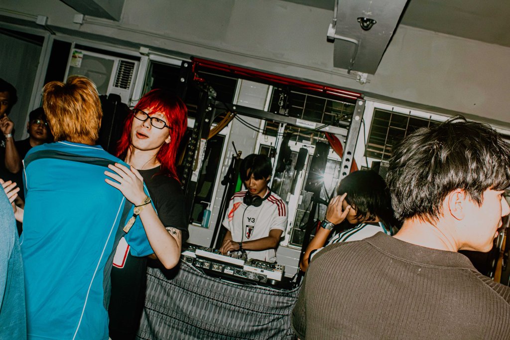 A lively scene at a music event with a DJ performing in the background. Several people are enjoying the atmosphere, with one person with red hair and glasses interacting with another in a blue shirt.