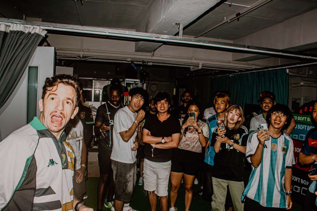 A diverse group of people gathered in a room, some cheering or reacting enthusiastically, wearing various soccer jerseys, with a mix of joyful expressions and smartphones in hand.