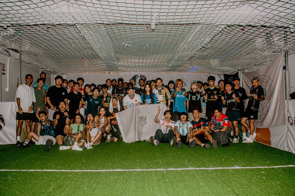 A large group of diverse individuals posing together indoors, celebrating in a sports environment with a netted ceiling and artificial grass floor.