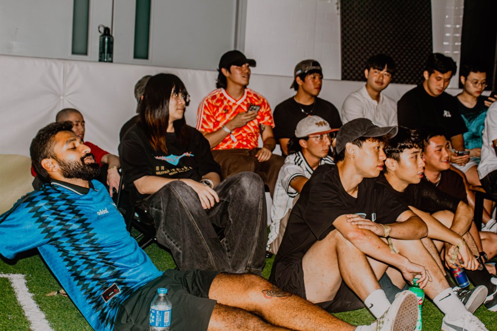 Group of young adults sitting together in a relaxed setting, some engaged in conversation while others are looking at their phones.