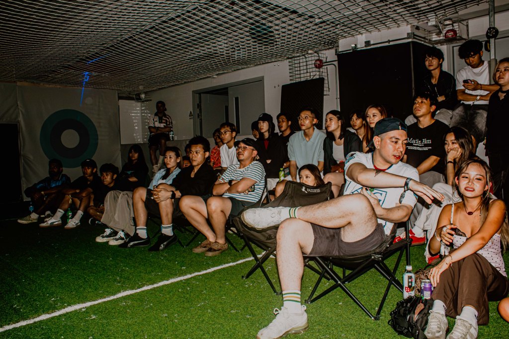 A diverse group of young people sitting on folding chairs in a dimly lit indoor space, looking engaged while watching something off-screen.