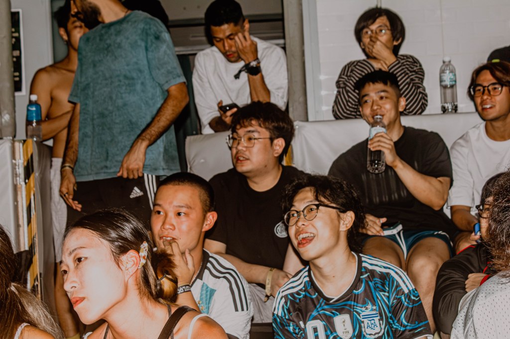 A diverse group of people enjoying an event, some displaying excited expressions, while others are focused on their devices. The setting appears lively and social.