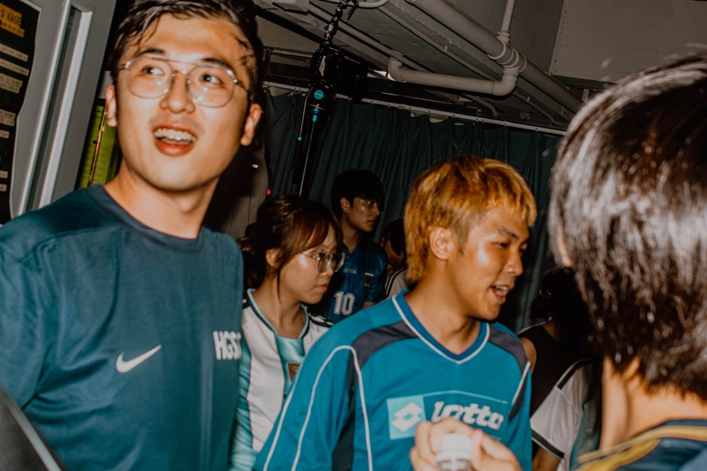 A group of young people in casual sports attire, socializing in a lively indoor setting. Two individuals in the foreground are engaged in conversation, with one wearing glasses and a blue T-shirt, while another has blonde hair and a blue sports jersey. The atmosphere appears energetic and friendly.