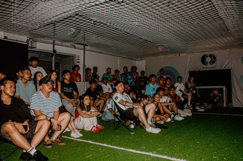 A diverse group of people seated in a sports facility, watching a presentation or event, with a grass-like floor and a soccer goal visible in the background.