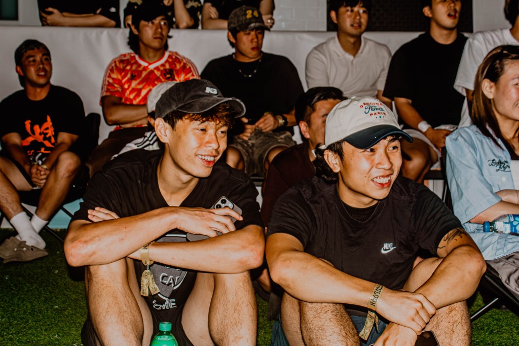 Two young men sitting closely together, smiling while watching an event, surrounded by an audience in casual attire.
