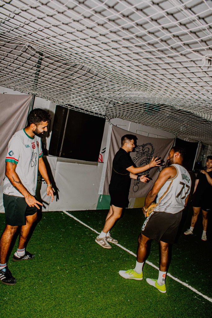 A group of four people engaged in a playful interaction in an indoor sports facility, with artificial turf and a netted ceiling.