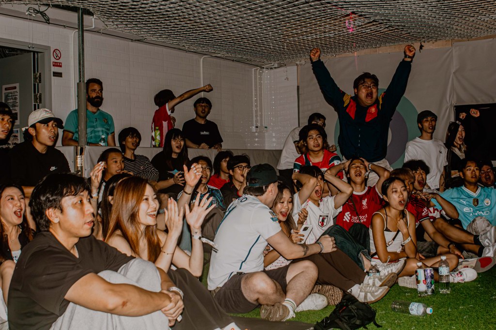 A lively crowd of diverse individuals watching a sports event, displaying a range of emotions from excitement to anticipation. Some are cheering and celebrating, while others are focused on the action, all gathered in a casual indoor setting.