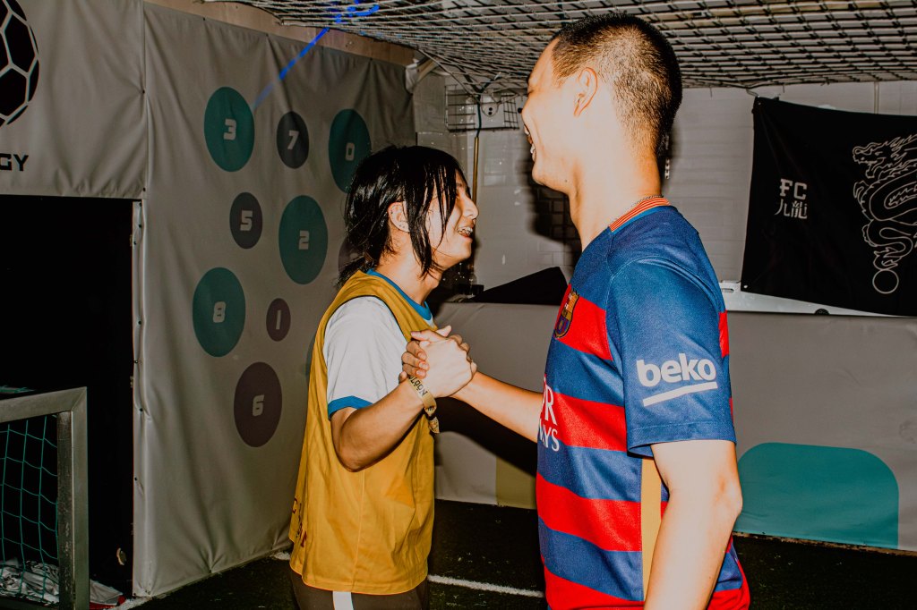 Two individuals smiling and shaking hands, one wearing a yellow sports jersey and the other in a blue and red striped soccer jersey, in a sports facility.