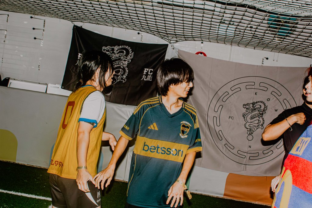 Two young players in a soccer training session, one wearing a yellow jersey and the other in a blue and gold jersey, with a sports-themed backdrop featuring flags and symbols.
