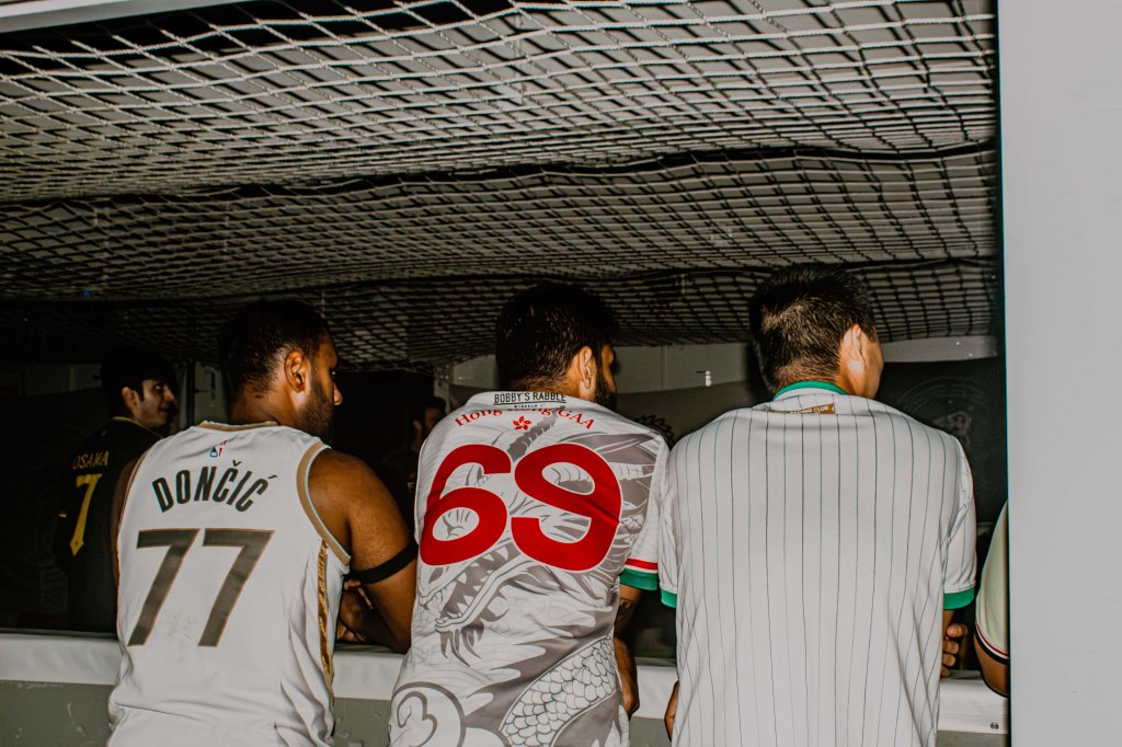 Three individuals wearing sports jerseys are seen from behind, looking out at a sports event. One jersey features the name 'DONTIC' with the number 77, another has the number 69, and the third has a striped design.